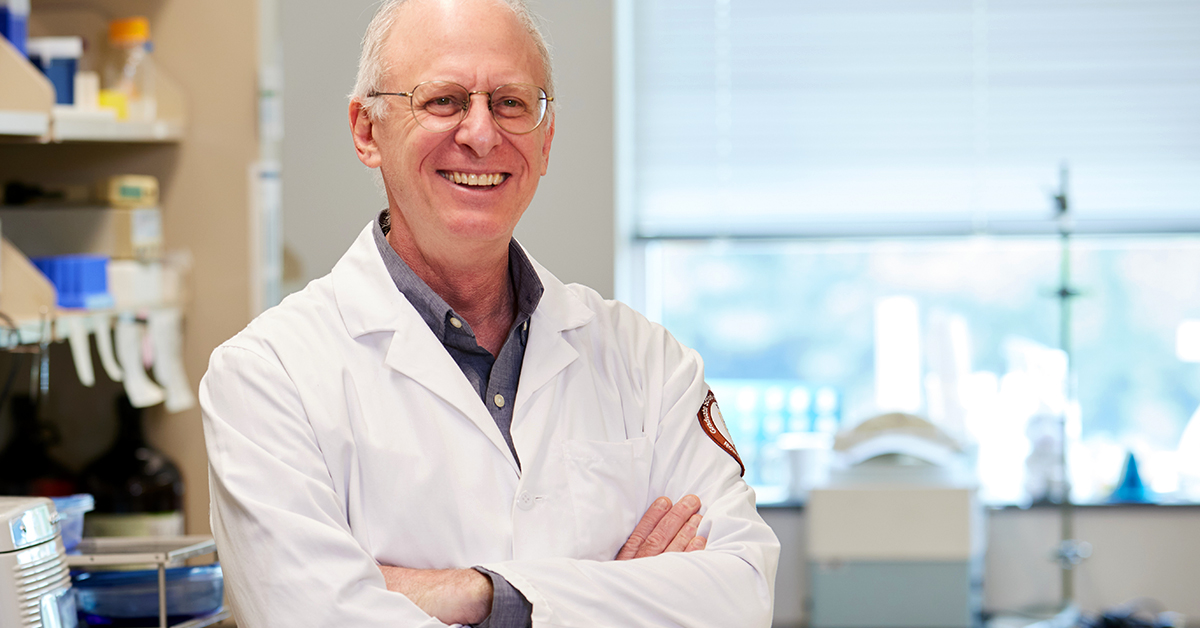 RowanSOM researcher begins human trials for cancer treatment drug. This is a portrait of Gary Goldberg, wearing a white lab coat. He has white hair and is smiling with his arms crossed in front of him. 