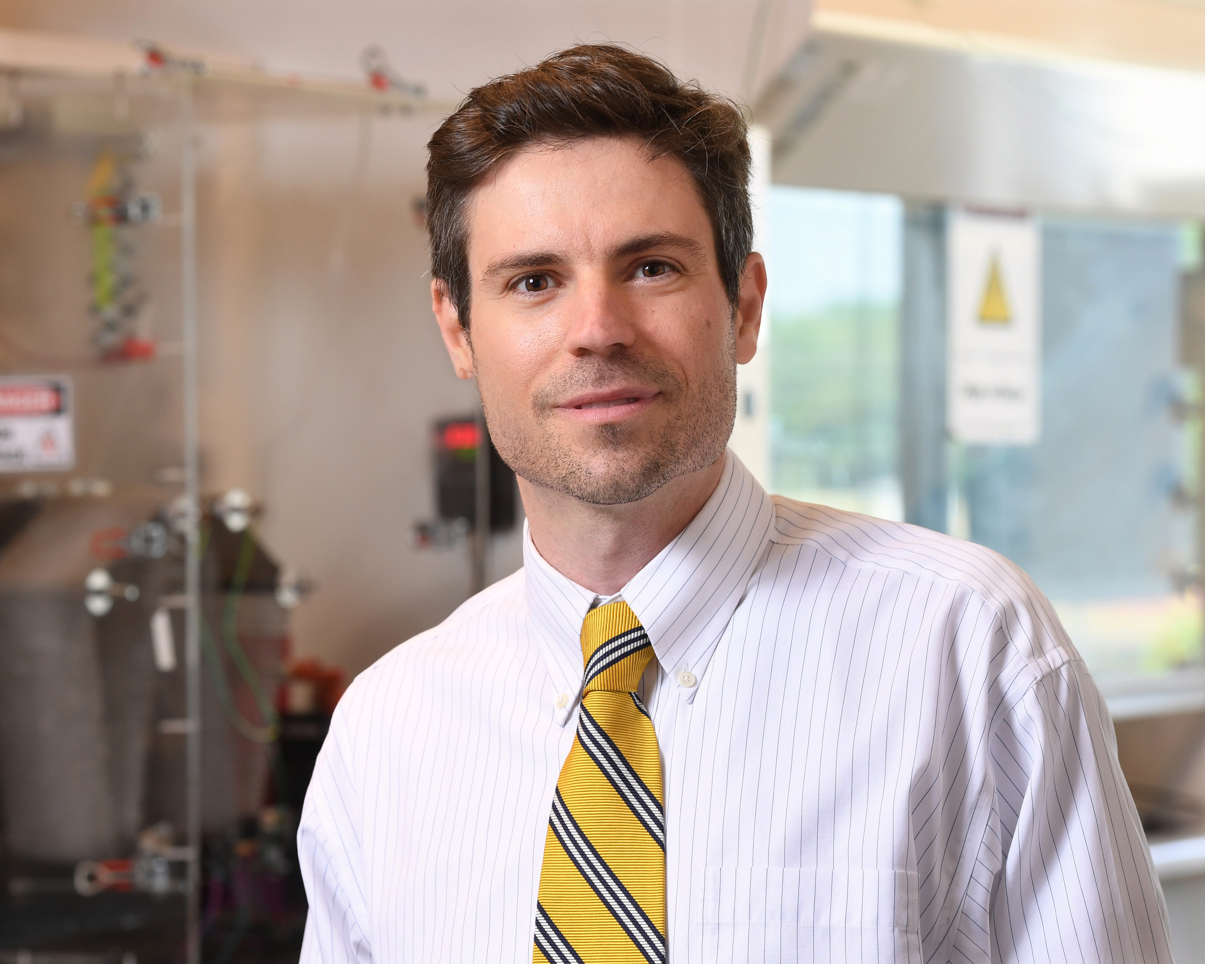 This is a portrait of Vincent Beachley, Ph.D., a white male with a slight smile, short brown hair touched with a little gray at the temples, wearing a white pinstriped dress shirt and yellow striped tie. He is standing in an engineering lab.