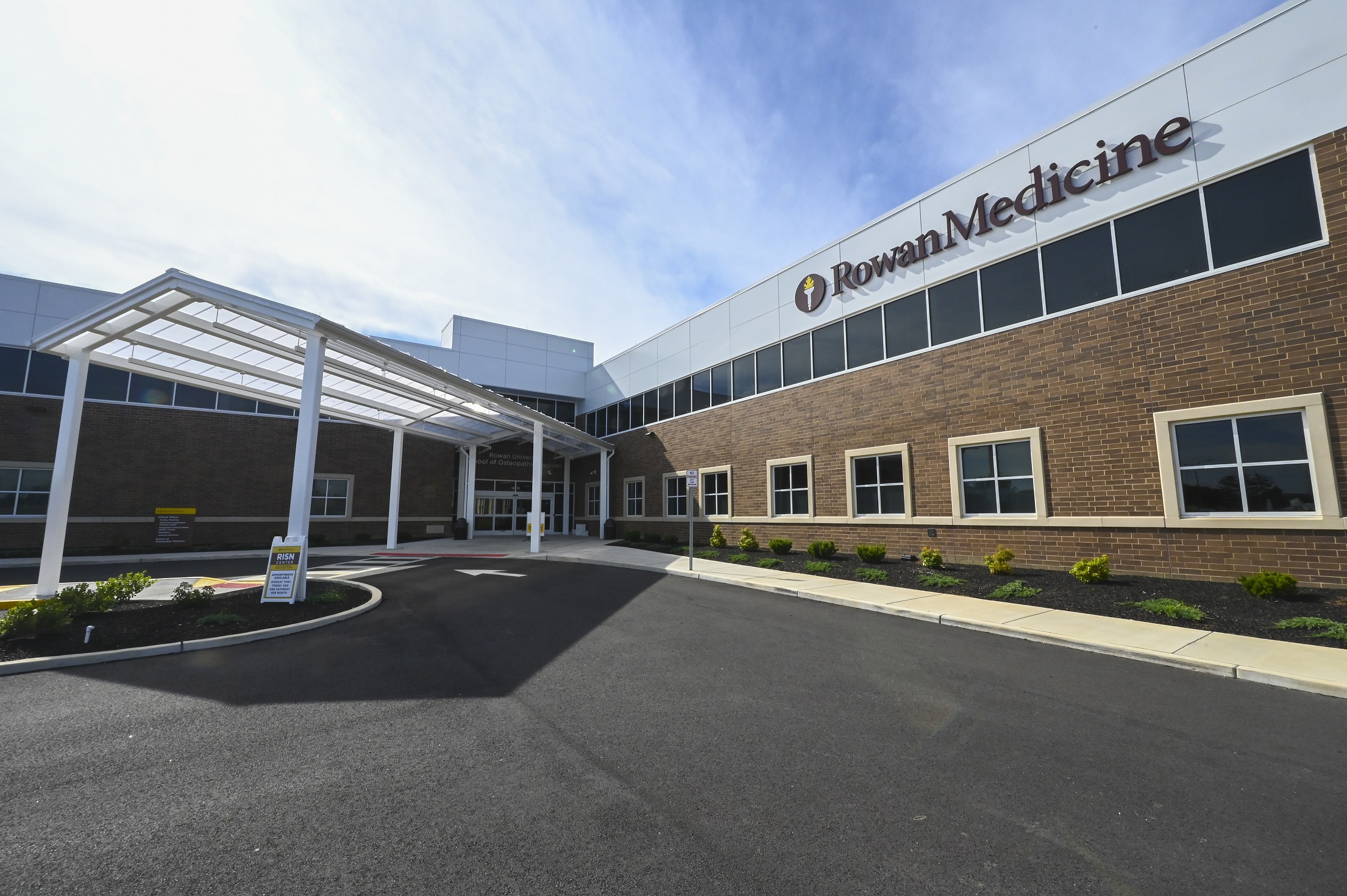 This is an exterior photo of Rowan Medicine in Sewell, N.J. It depicts the entrance to a brick two-story building with a frosted-glass awning shading the entrance and a circular driveway.