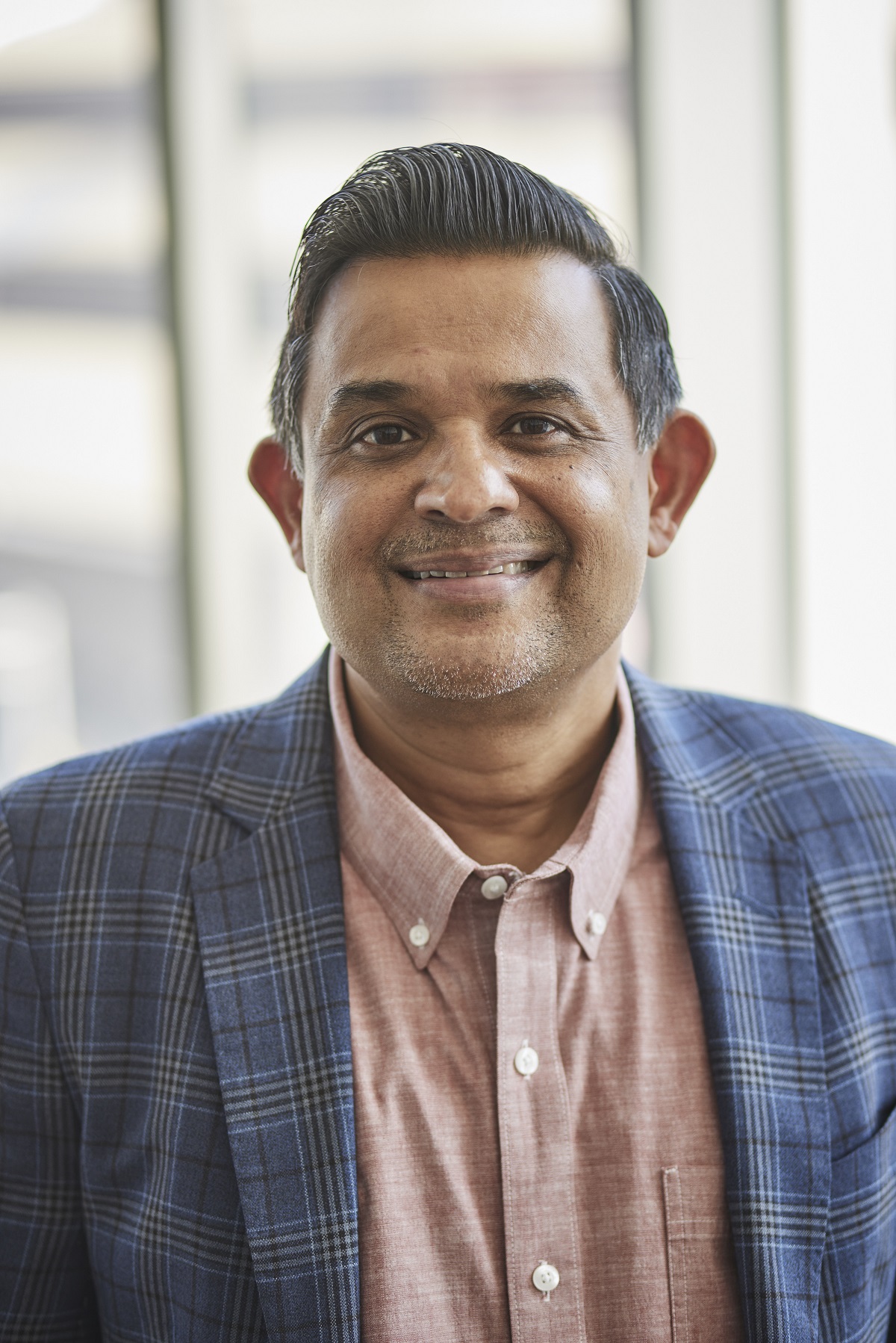 This is a portrait of Dr. Shreekanth Mandayam. He is standing in the Virtual Reality Center in Camden, wearing a blue plaid suit jacket and an open-necked dusky rose buttoned dress shirt. He is a middle-aged man with medium brown skin, black hair and brown eyes. He is relaxed and smiling at the camera.  