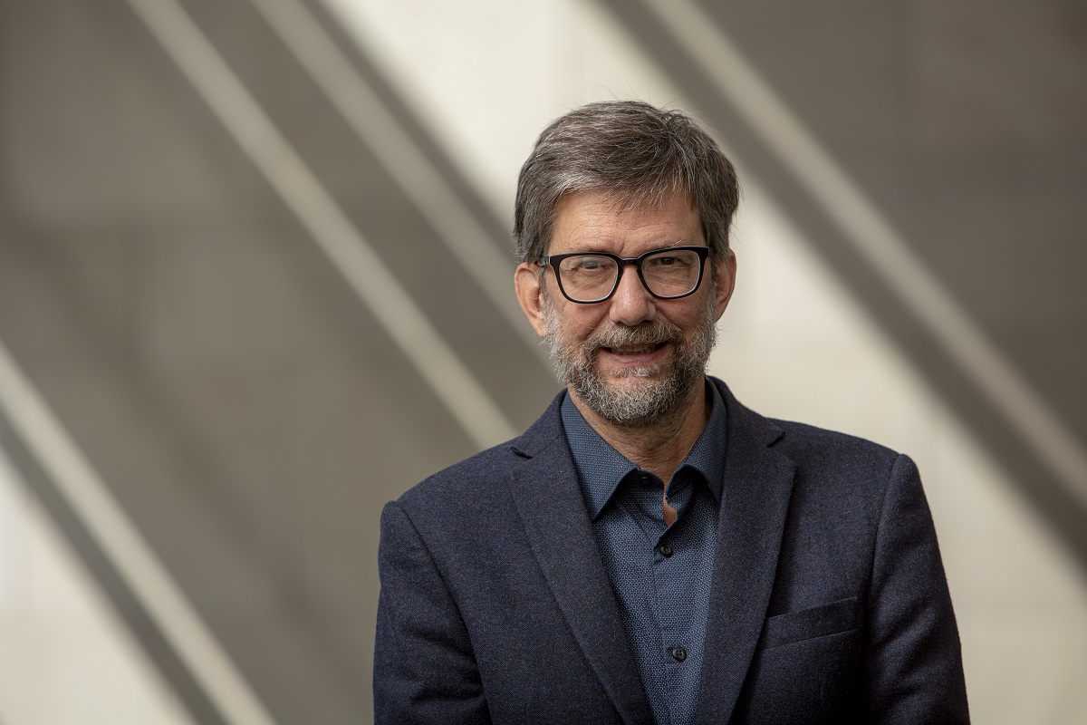 This is a portrait of Dr. Jess Everett, a middle-aged white male who is smiling slightly at the camera. He's wearing a navy wool suit jacket and a navy blue dress shirt. He has black-rimmed glasses and a medium-length, salt-and-pepper beard. 