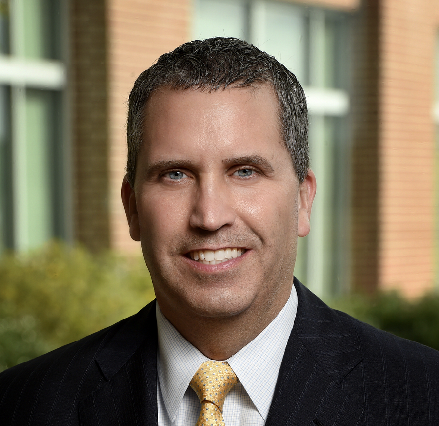 This is a professional portrait of Mark Byrne, Ph.D. He is a middle-aged white man with short salt-and-pepper hair and blue eyes. He is smiling and is wearing a dark suit jacket, white shirt and yellow tie