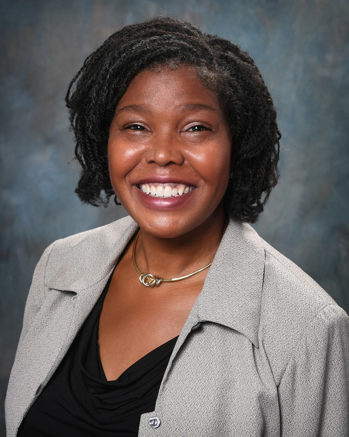 This is a portrait of Dr. Tabbetha Dobbins, a Black woman with dark curly hair threaded with silver strands. She is smiling and wearing a tweed suit jacket over a dark blouse. 
