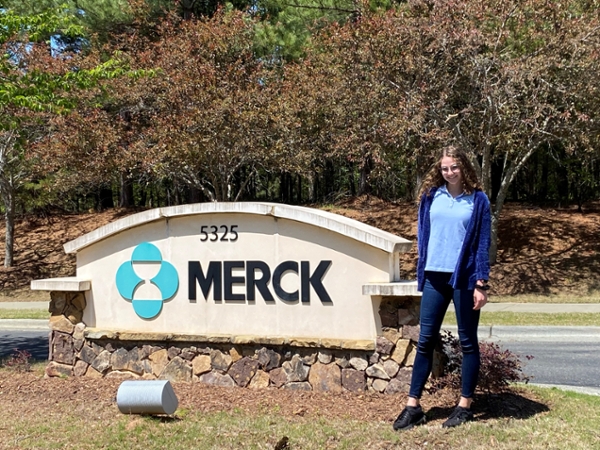 Lauren Repmann stands in front of an exterior sign displaying the name, Merck. She is a young adult female with long curly, blonde hair. She is wearing glasses and a blue polo shirt with a cardigan and slim-fitting pants. 