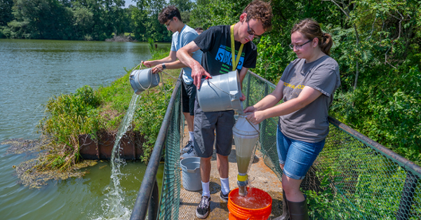 EDGe Lab students collect water samples on a pedestrian bridge above a small dam on a lake.