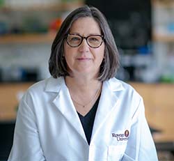 This is a portrait of Alison Krufka, PhD. She is wearing a lab coat and glasses, and looking into the camera. She is a white woman with straight, shoulder-length salt-and-pepper hair. 