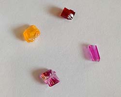A close-up shot of four crystals with different geometric shapes and dyed red, pink and yellow sit on a white background.
