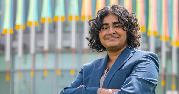 Kavin Haldo wears a blue suit and stands confidently with crossed arms against a blurred background of colorful vertical lines.