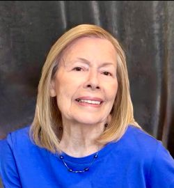 A formal portrait shows a smiling woman with straight blonde shoulder-length hair, wearing a blue top and beaded necklace against a gray fabric backdrop.