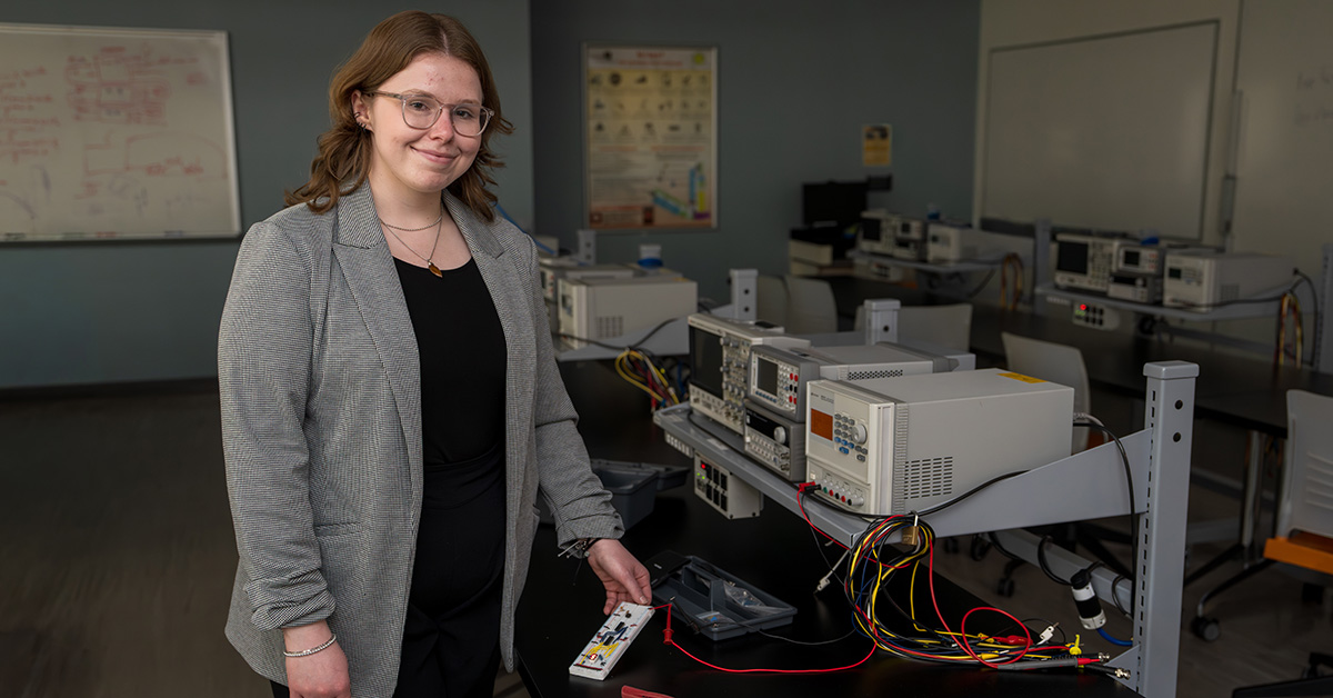A young woman wearing a business suit stands in an empty electrical engineering classroom while holding a small electrical module.