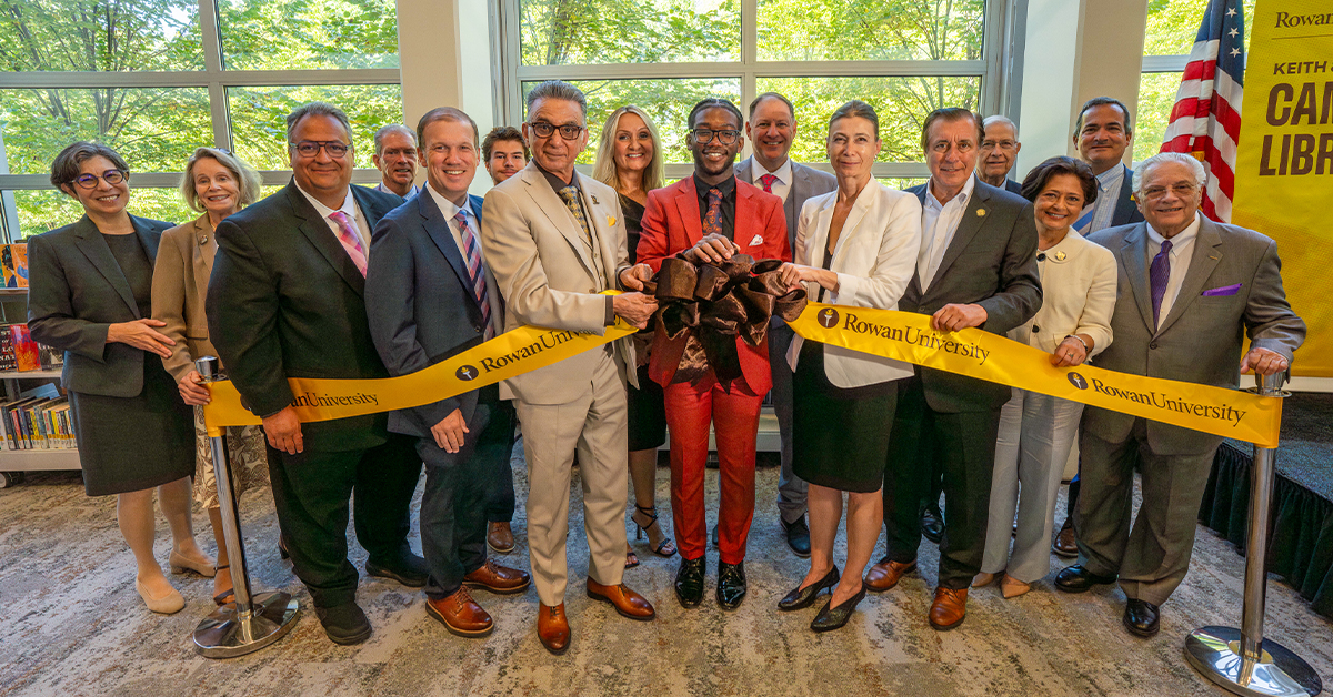 A group of administrators, friends and board members hold a long yellow ribbon in a crowded room and smile at the camera during a crowded event at the library. 