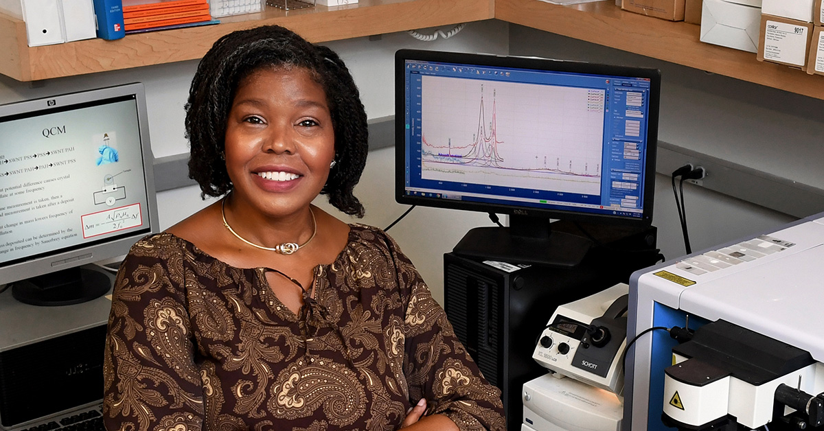 A woman wearing business attire sits in a lab in front of a computer screen and lab equipment.